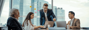 Four business professionals collaborating around a laptop in a high-rise conference room with a city skyline backdrop and colorful sticky notes on the window.